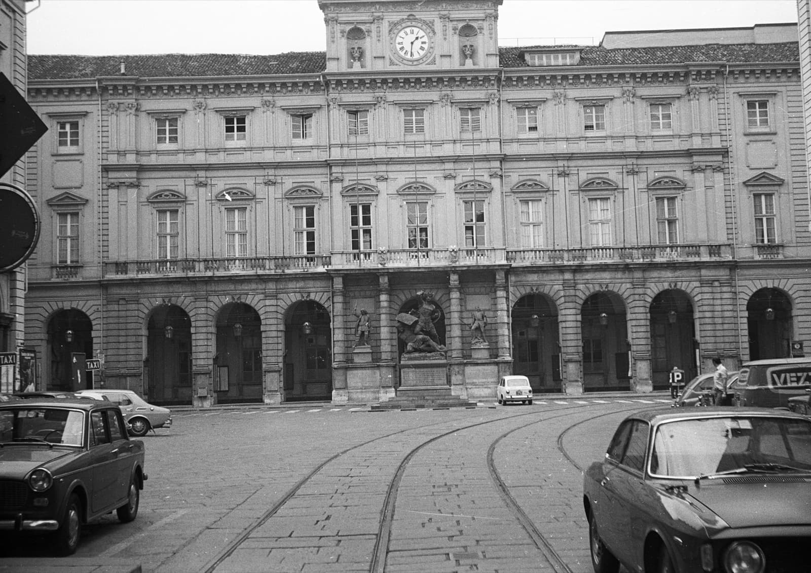 Black-and-white photograph of an arcaded town hall with a clock, small cars and tram rails in the square