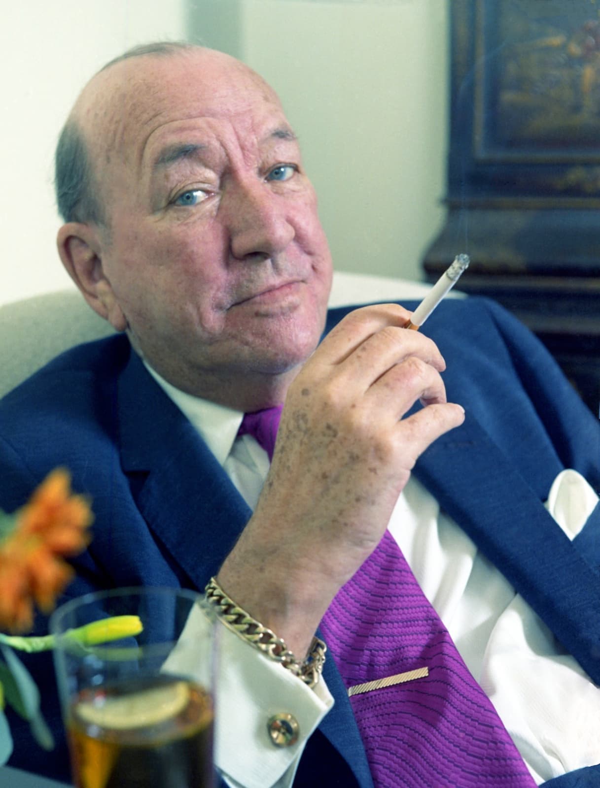 Colour portrait of Noël Coward in a blue suit and purple tie, cigarette in hand