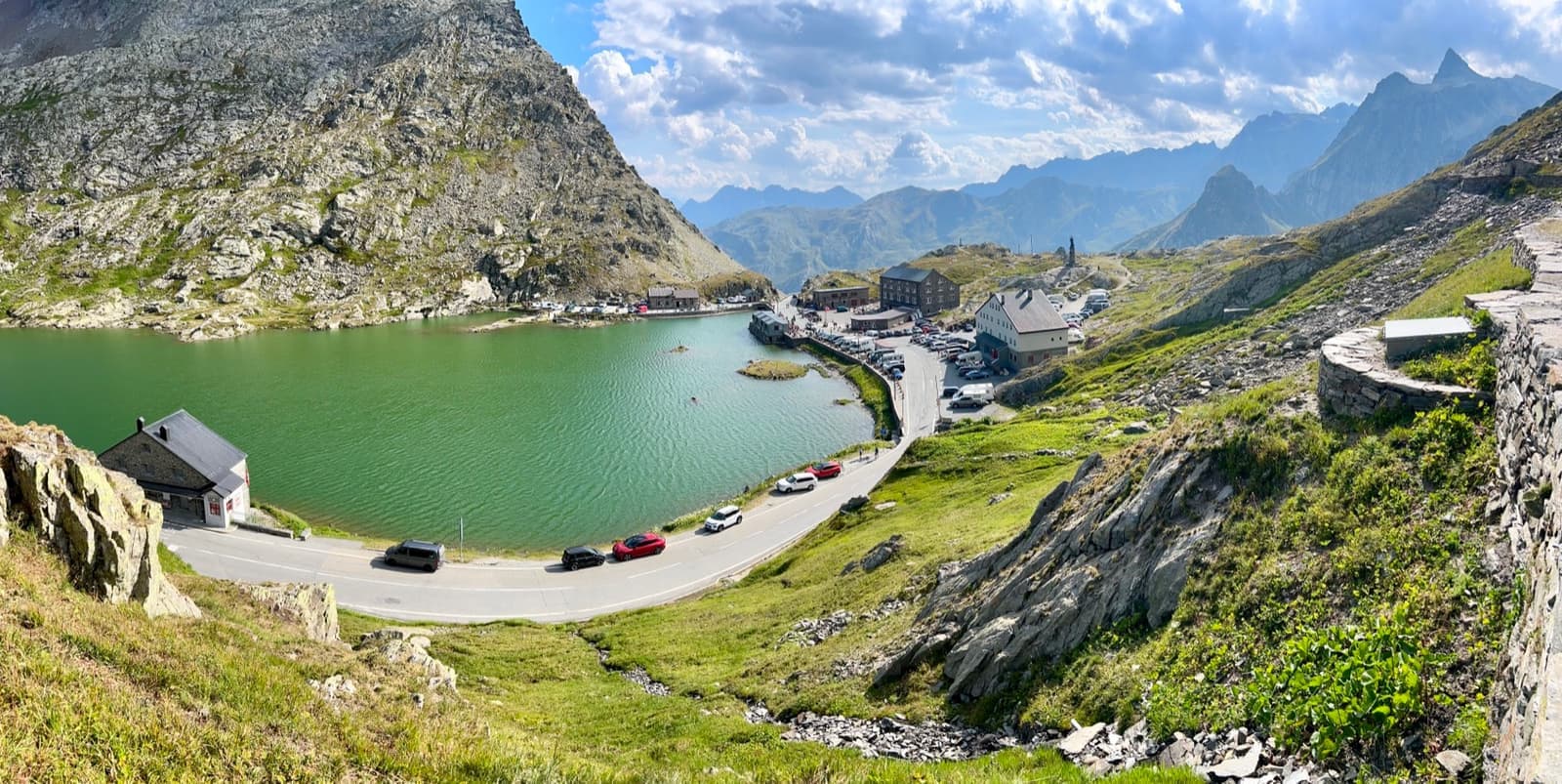 A green mountain lake below a road and a hospice building, with rocky peaks and blue sky