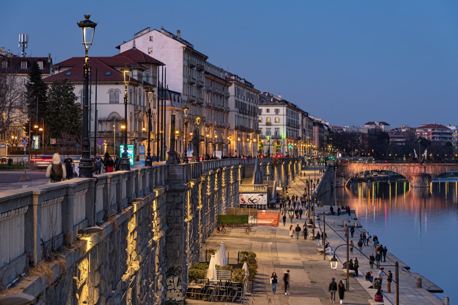 A stone river embankment with arches at water level and lamplit buildings above, at dusk