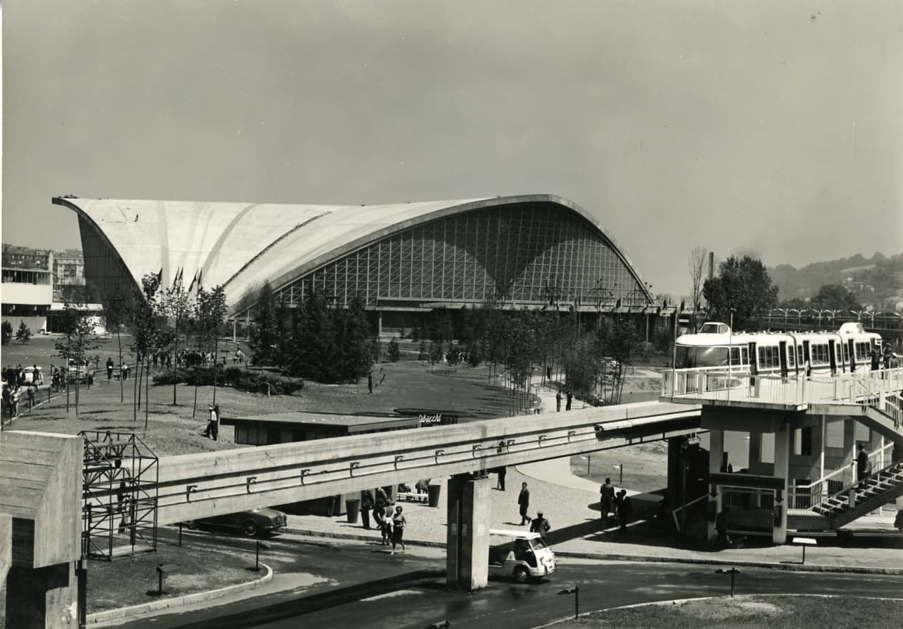 Black-and-white photograph of a building with a huge curved sail-shaped concrete roof and a walkway in front