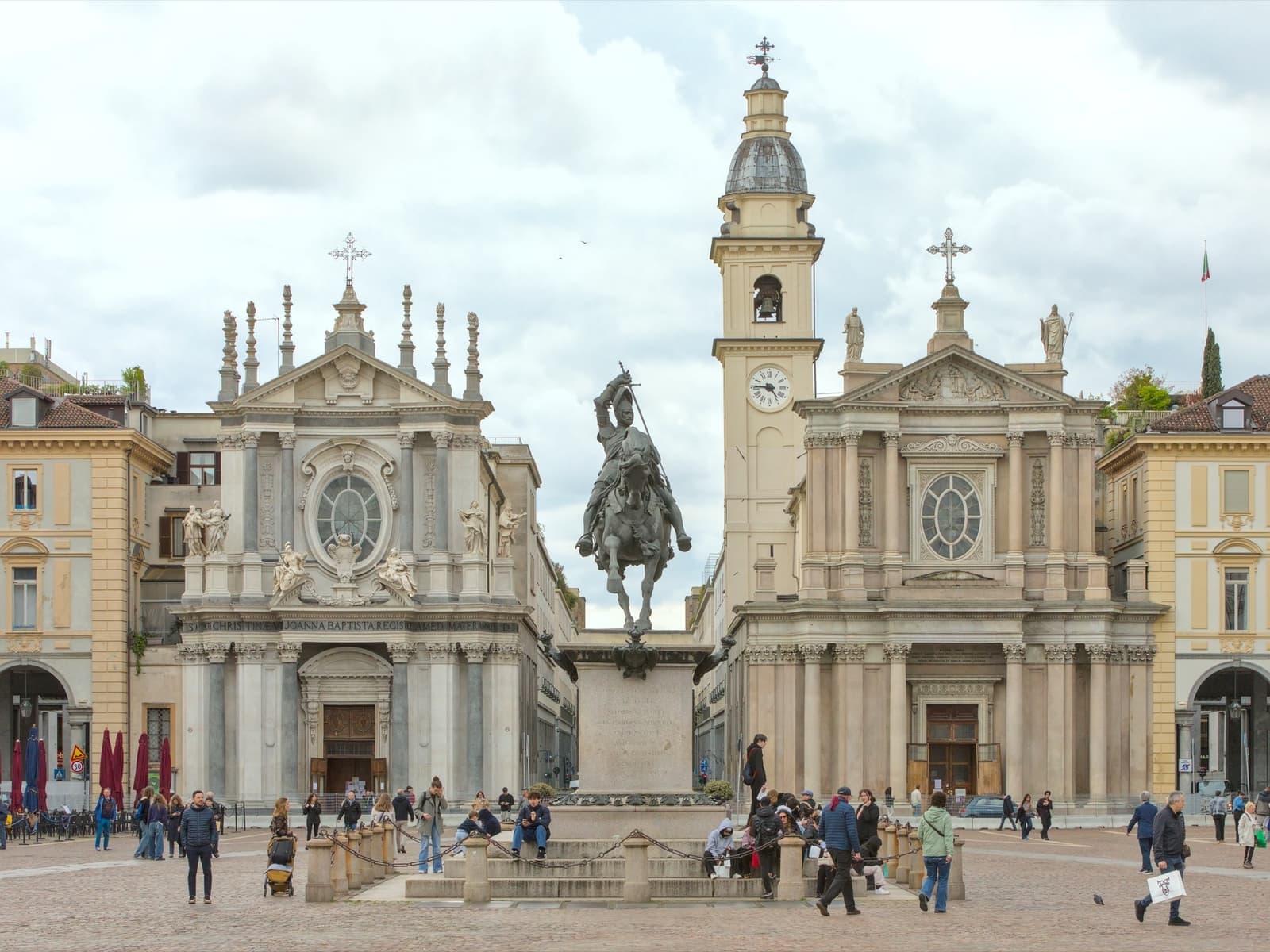 A grand square with twin baroque churches and an equestrian statue, people walking