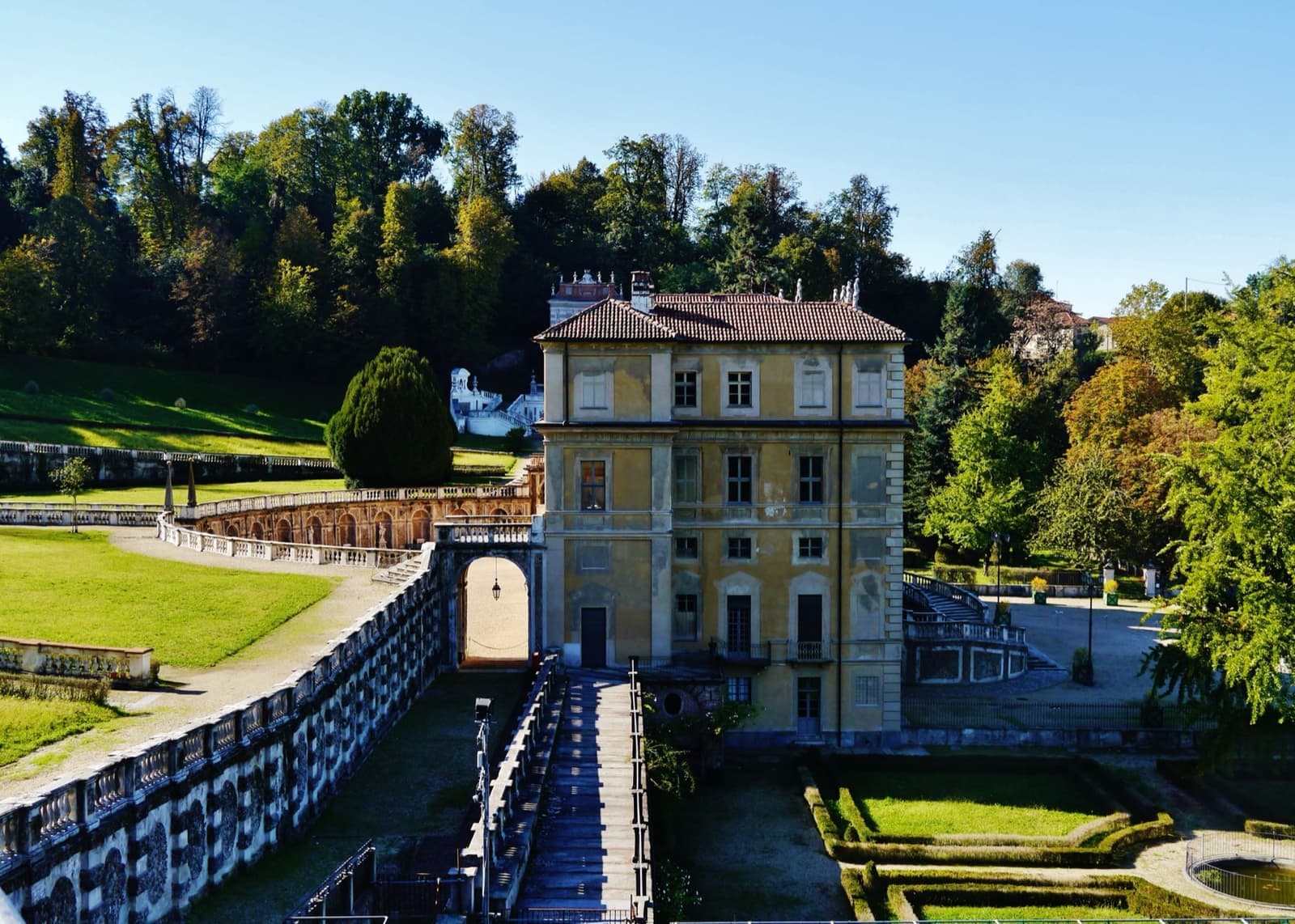 A pale yellow villa among trees with terraced lawns and a balustrade below