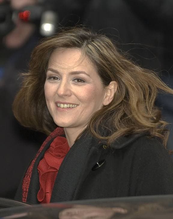 Martina Gedeck smiling, hair blown back, in a dark coat and red scarf