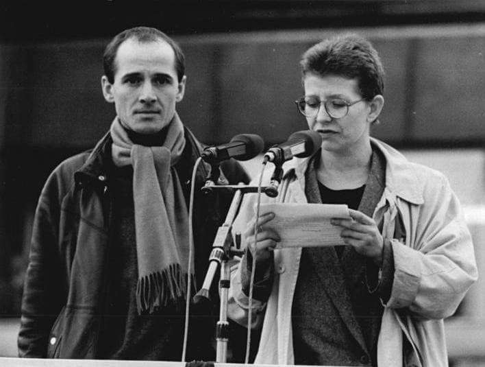 Ulrich Mühe in a scarf beside Johanna Schall, who reads from a sheet at a microphone