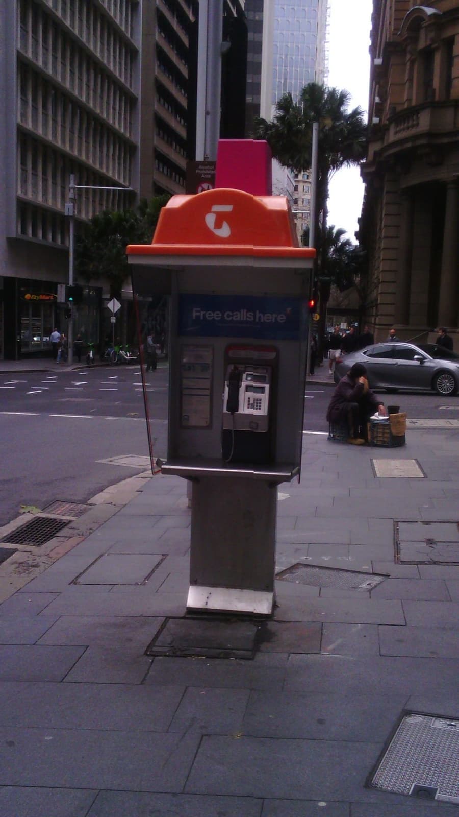A modern orange-hooded payphone on a paved corner between office towers