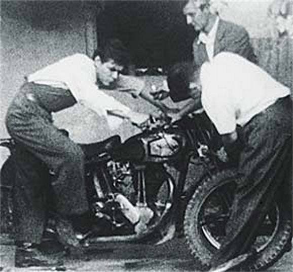 A young man leans over the handlebars of an old motorcycle while two men bend over the engine
