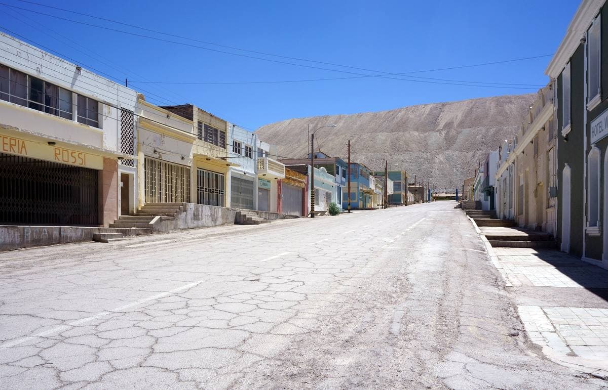 A wide empty street of low shuttered buildings below a grey spoil heap