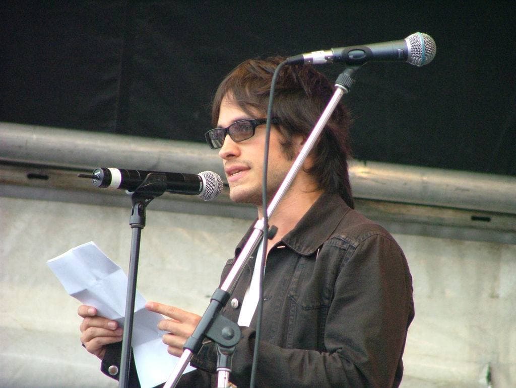 Gael García Bernal in glasses and a dark jacket, reading from a sheet at a microphone
