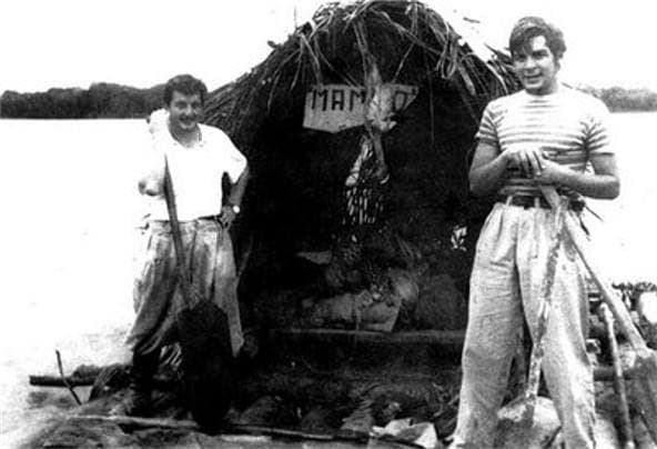 Two young men stand either side of a palm-thatched shelter on a raft, a sign reading MAMBO on it