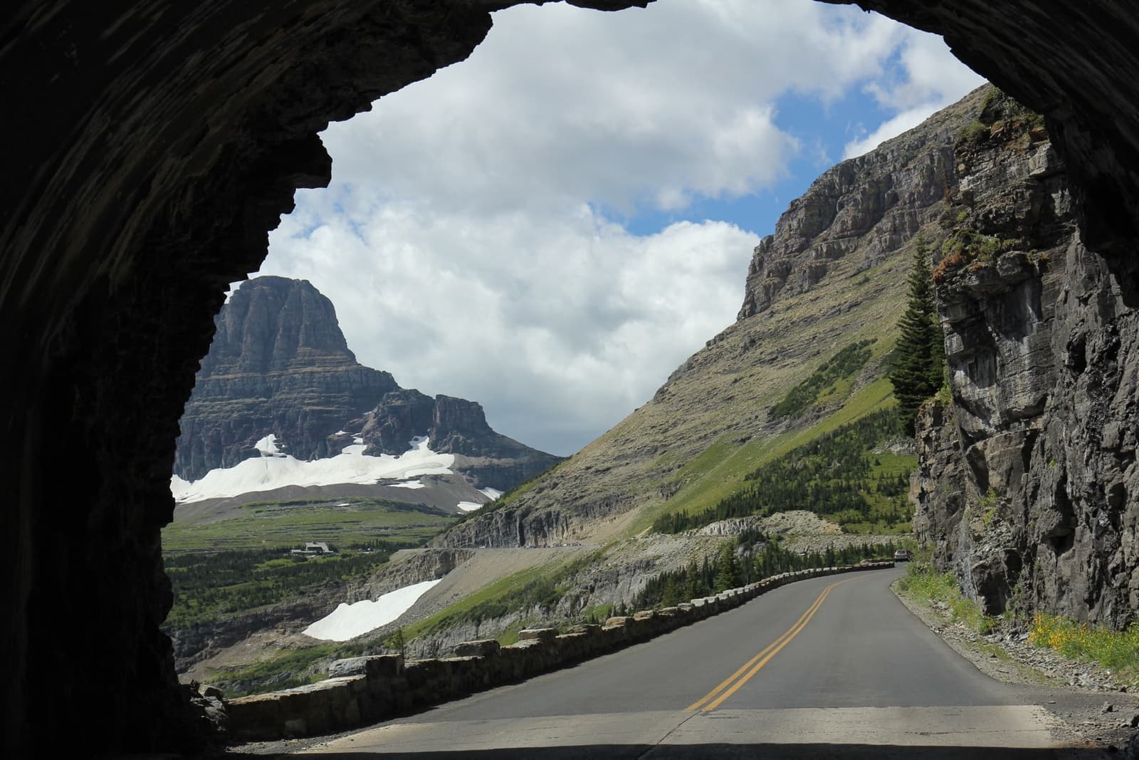 A road curving out of a rock tunnel along a mountainside towards snowy peaks