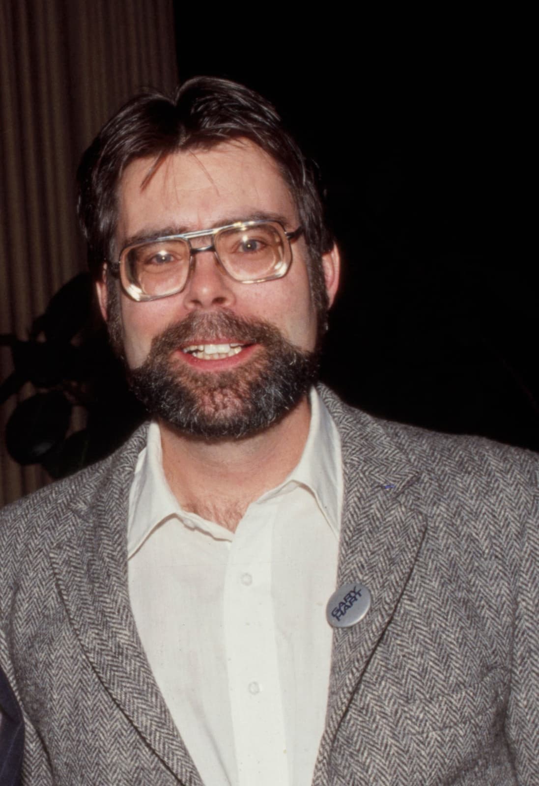 Stephen King with a beard and glasses, in a tweed jacket, smiling
