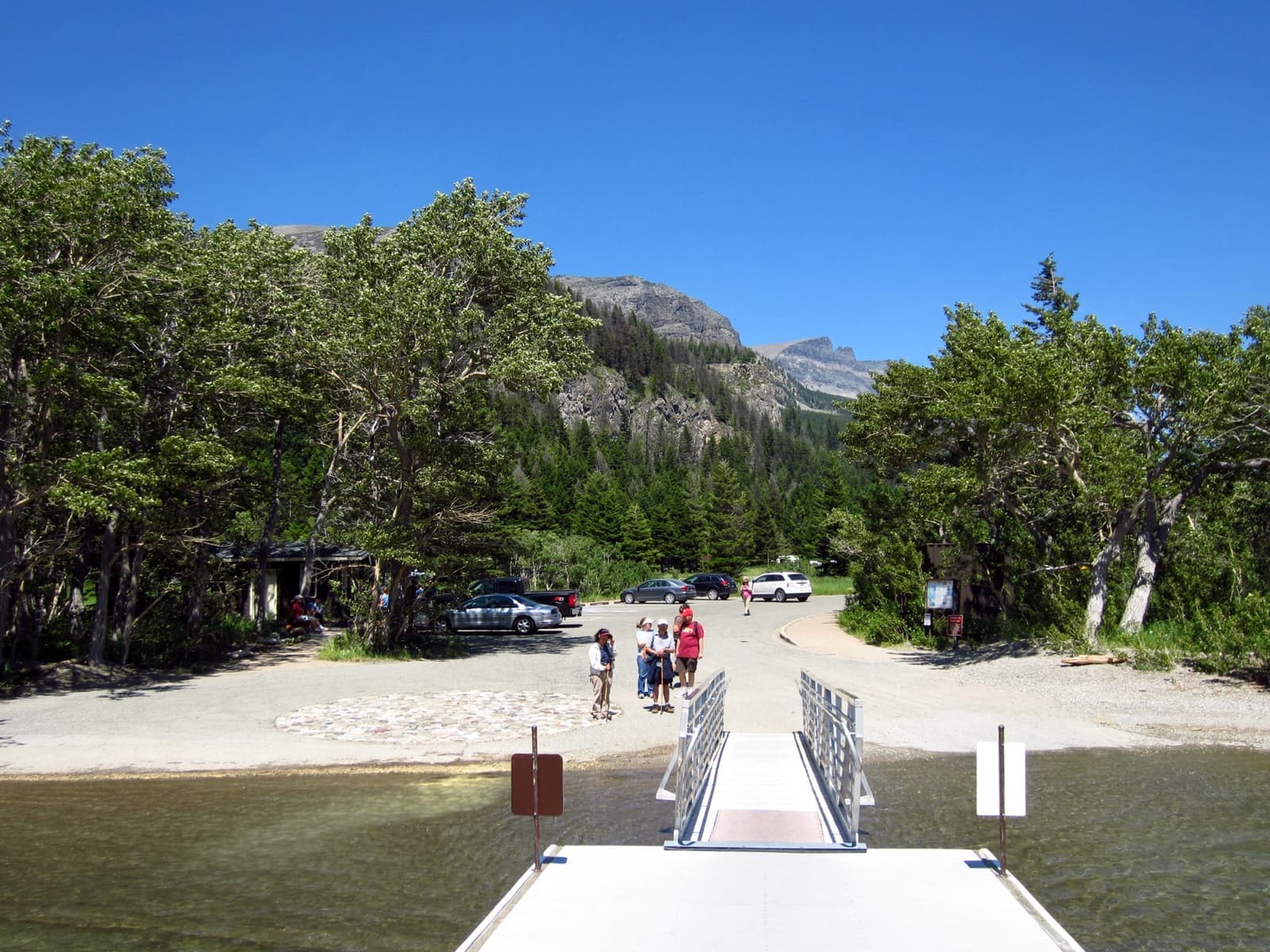 A white jetty running from a pebble beach into clear water below wooded slopes