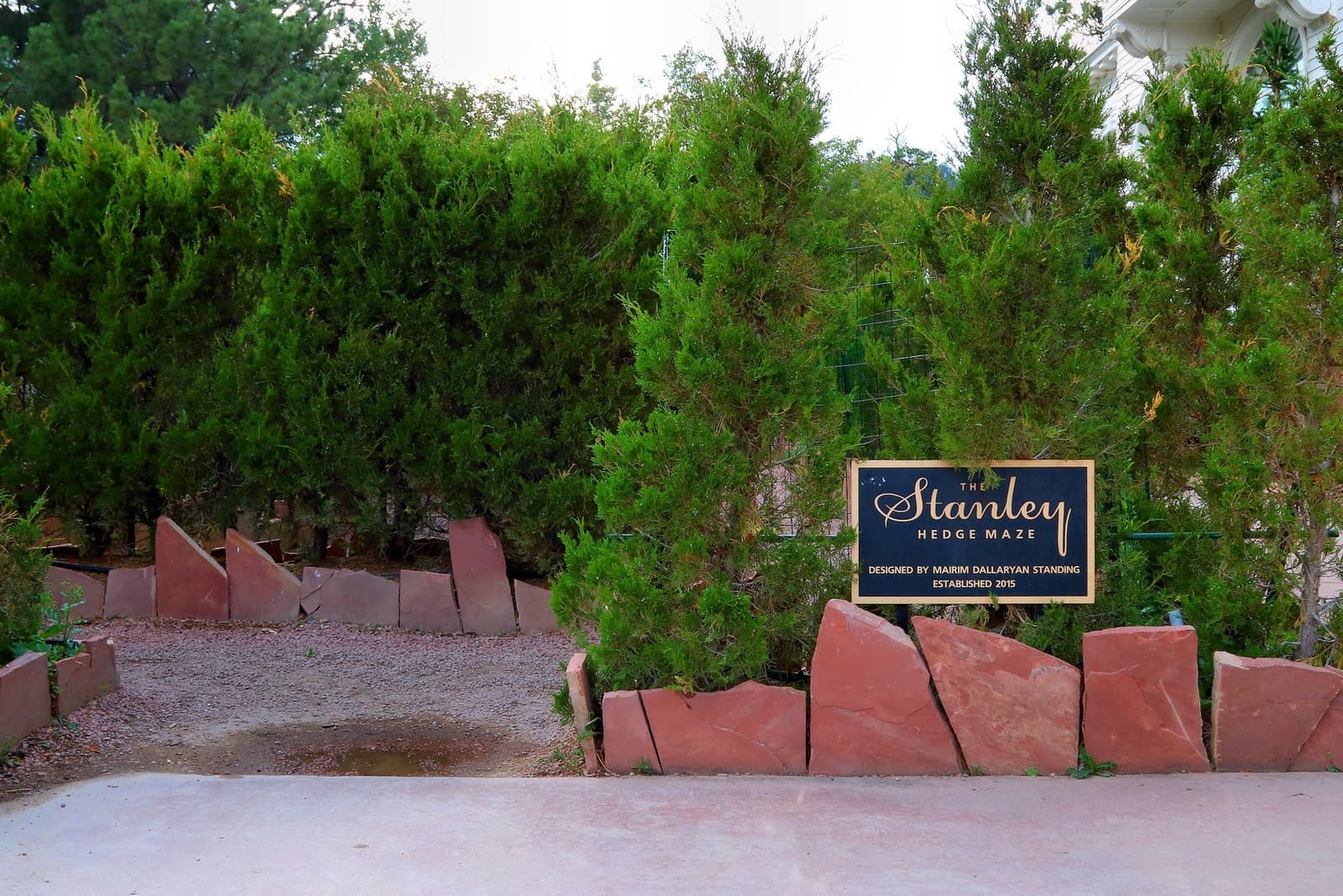 A gap in a green juniper hedge behind red stone edging and a small sign