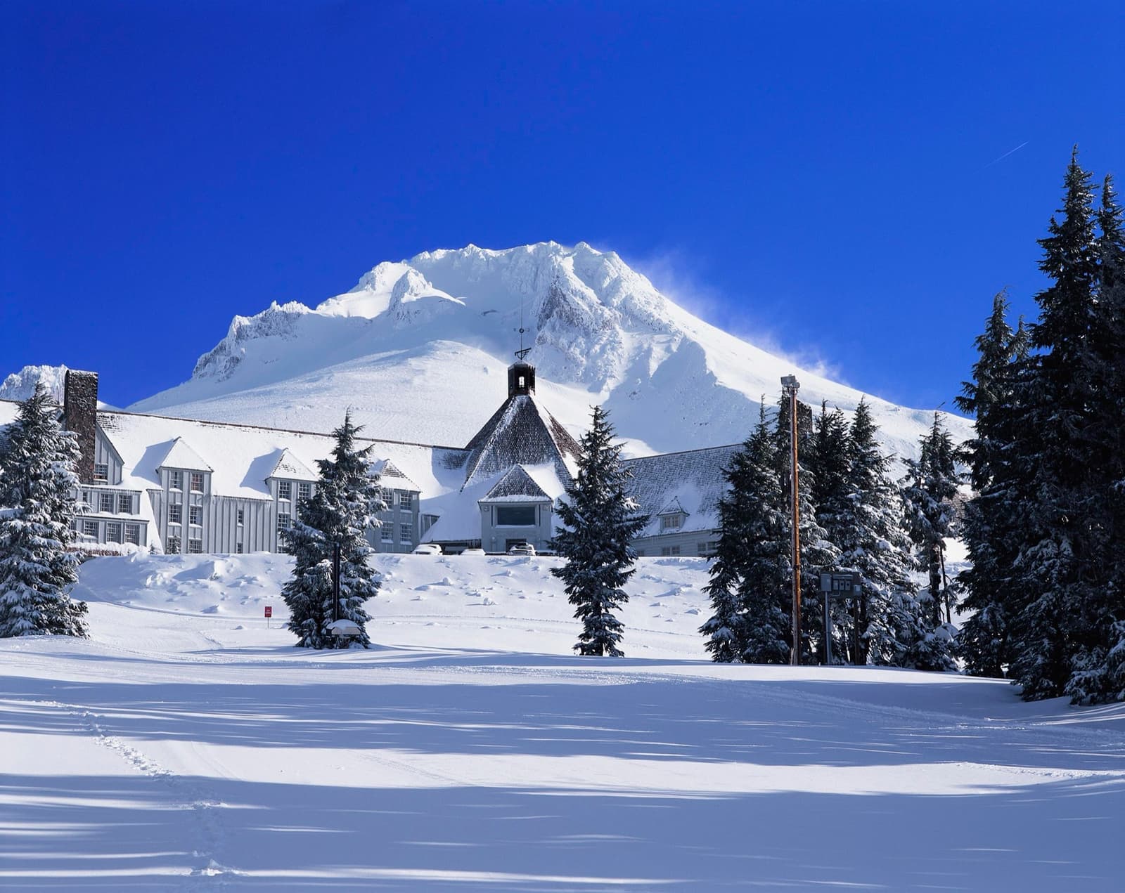 A long timber lodge with a steep roof half buried in snow below a white mountain