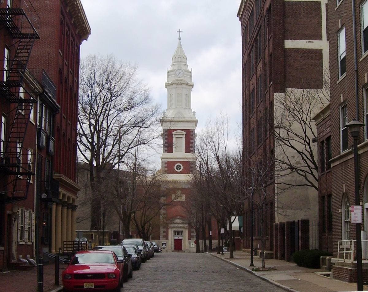 A narrow Old City street lined with parked cars and brick buildings, a church steeple at its end