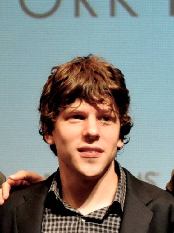 Jesse Eisenberg in a checked shirt and dark jacket, curly hair, looking at the camera