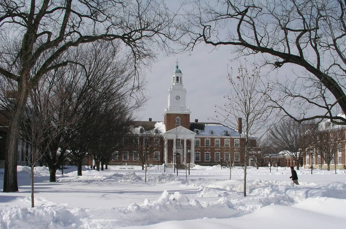 A brick hall with a white clock tower across a snow-covered quad