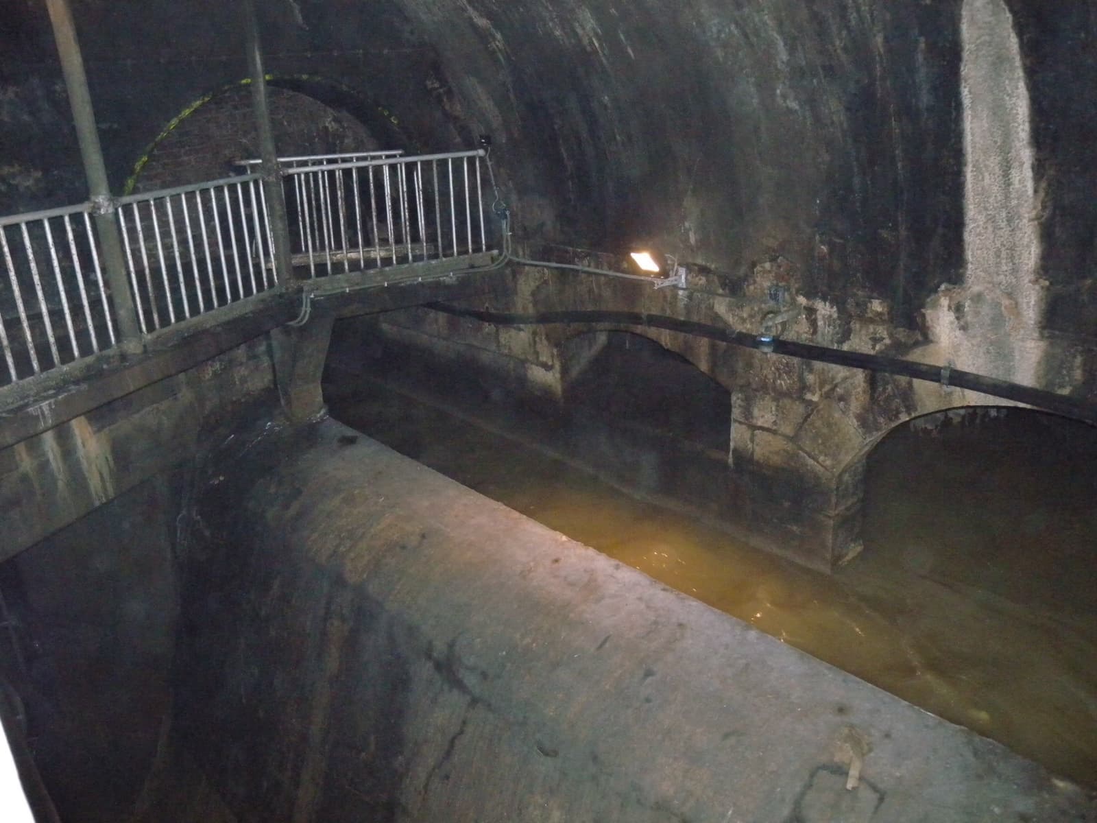 A vaulted brick sewer channel with a railed walkway, lit by lamps