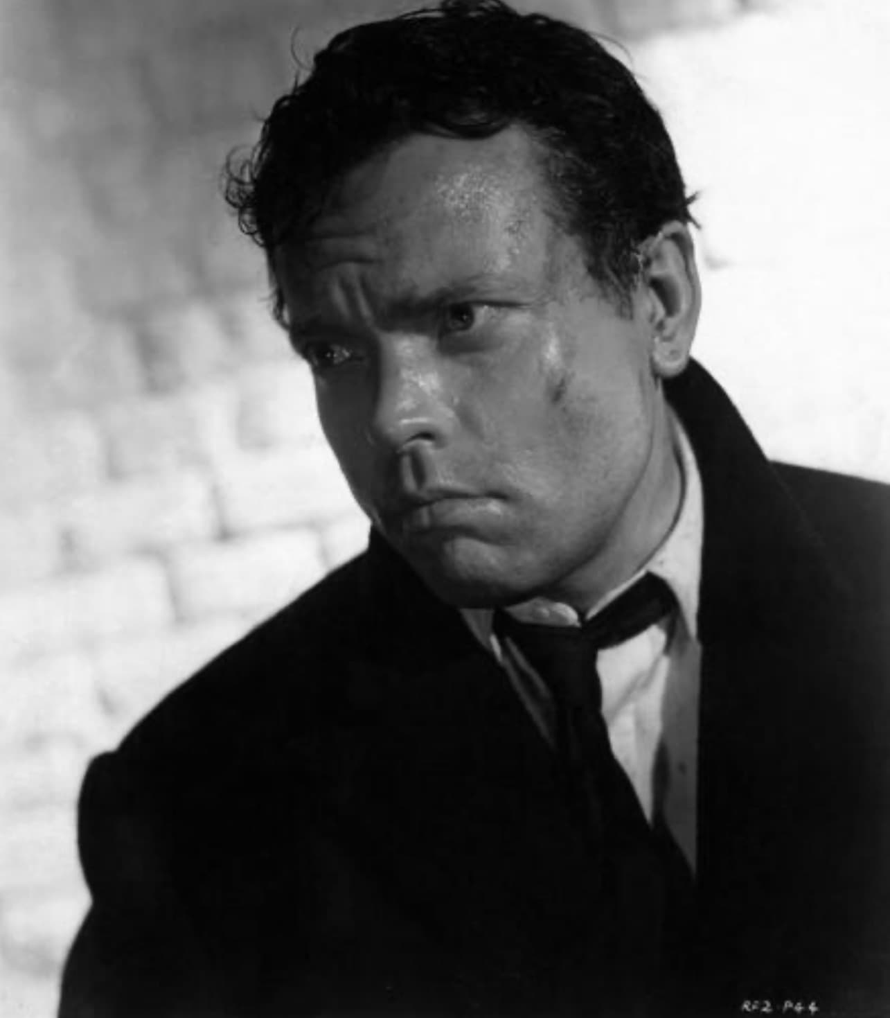 Orson Welles in a dark coat, looking sideways against a pale wall