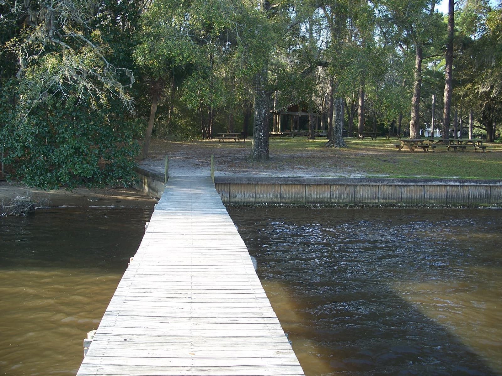 A long weathered wooden dock running out over brown bayou water, with oaks and lawn on the shore