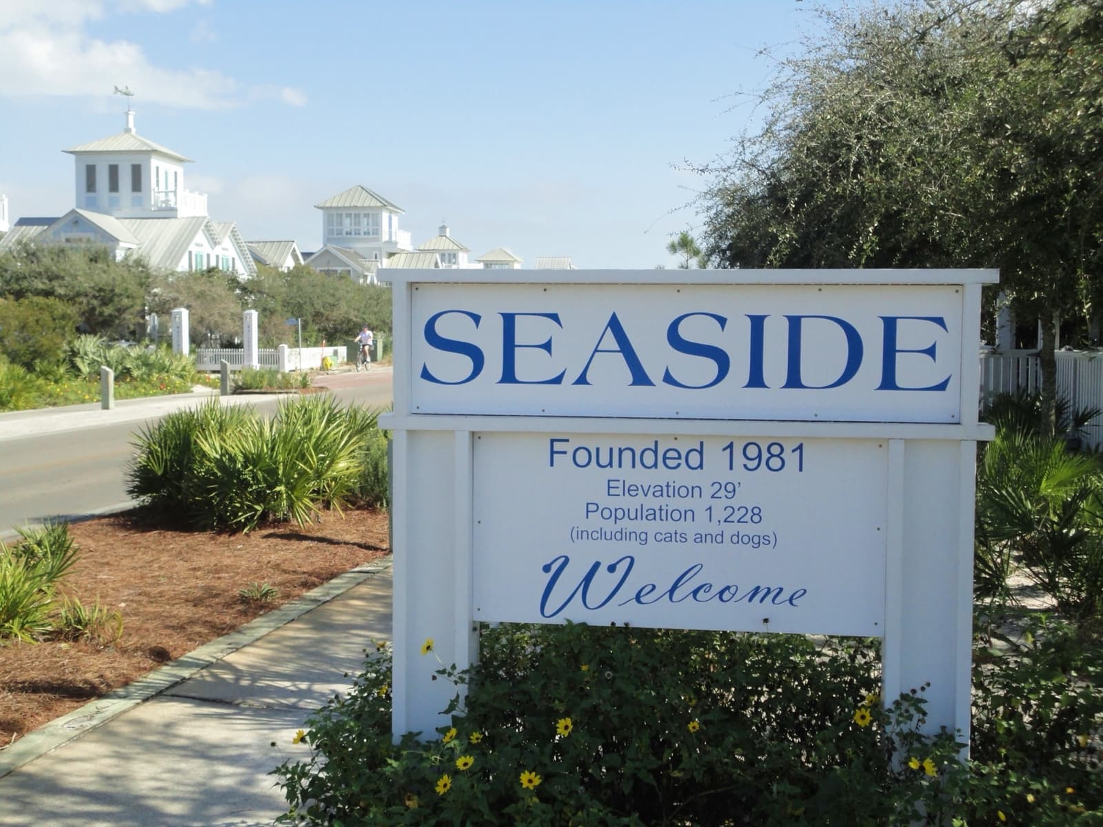 A white sign reading Seaside, Founded 1981, Welcome, with white houses and towers behind