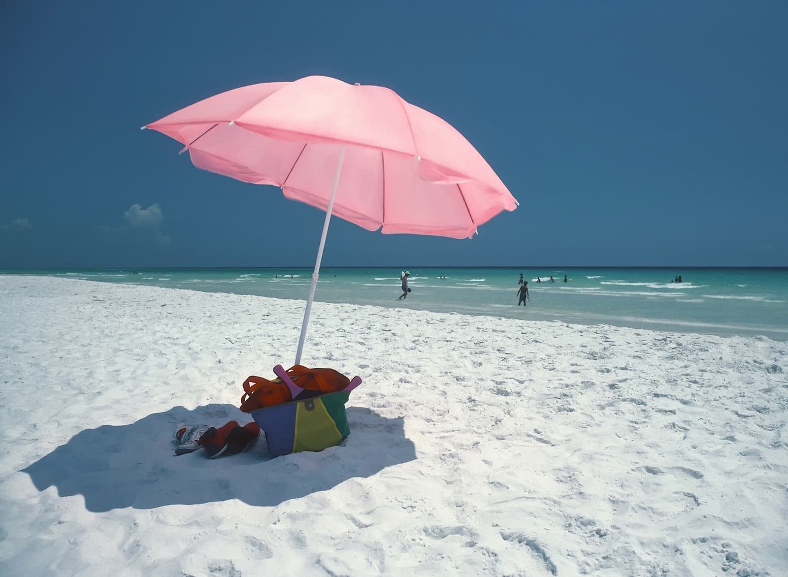 A pink umbrella on white sand, with swimmers in turquoise water beyond