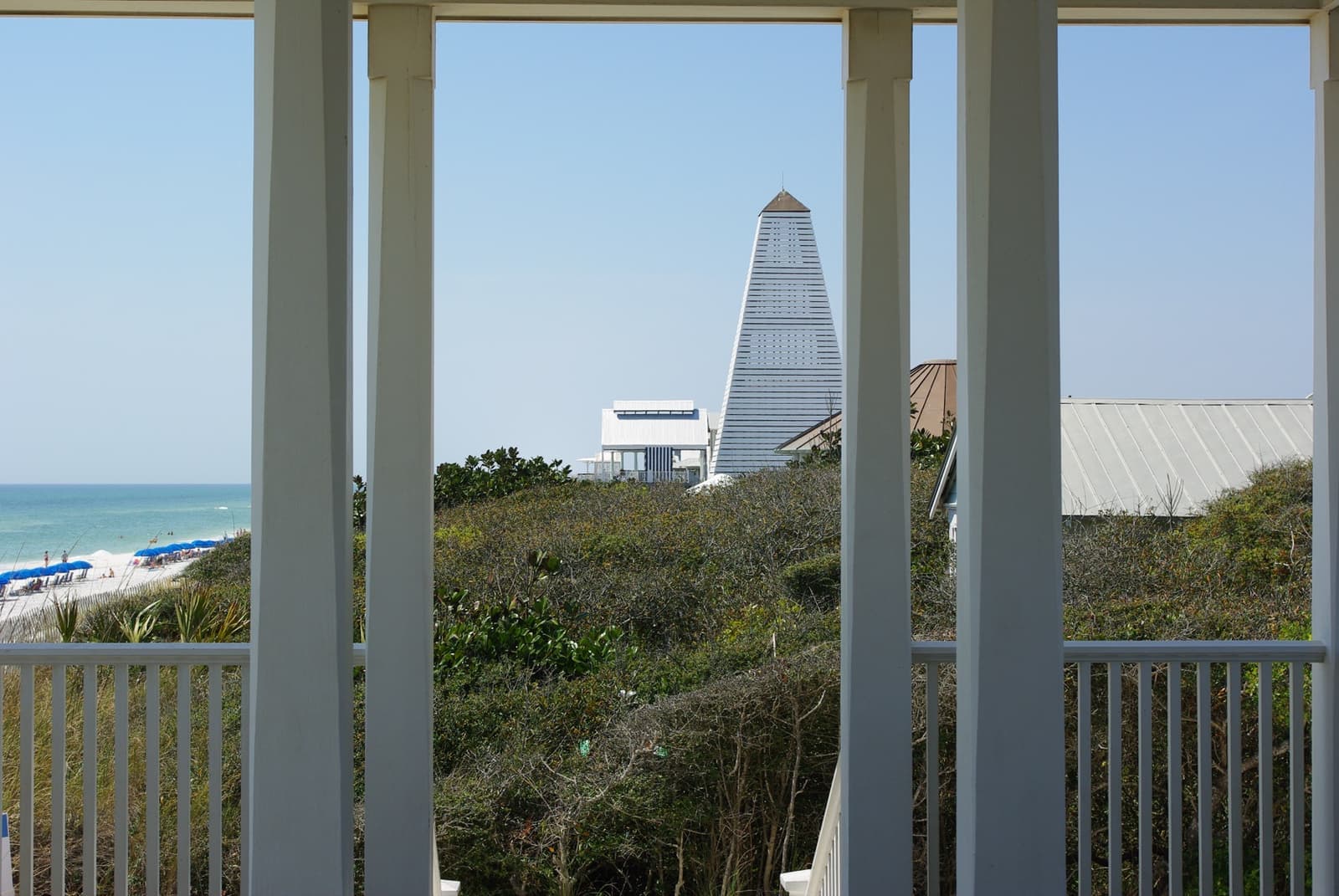 Through white porch columns, a tall pointed beach tower stands above the dunes and the blue Gulf