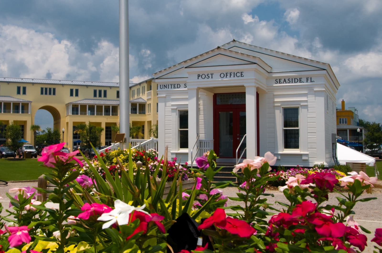 A small white temple-fronted post office with a red door, pink flowers in front and an arcaded building behind