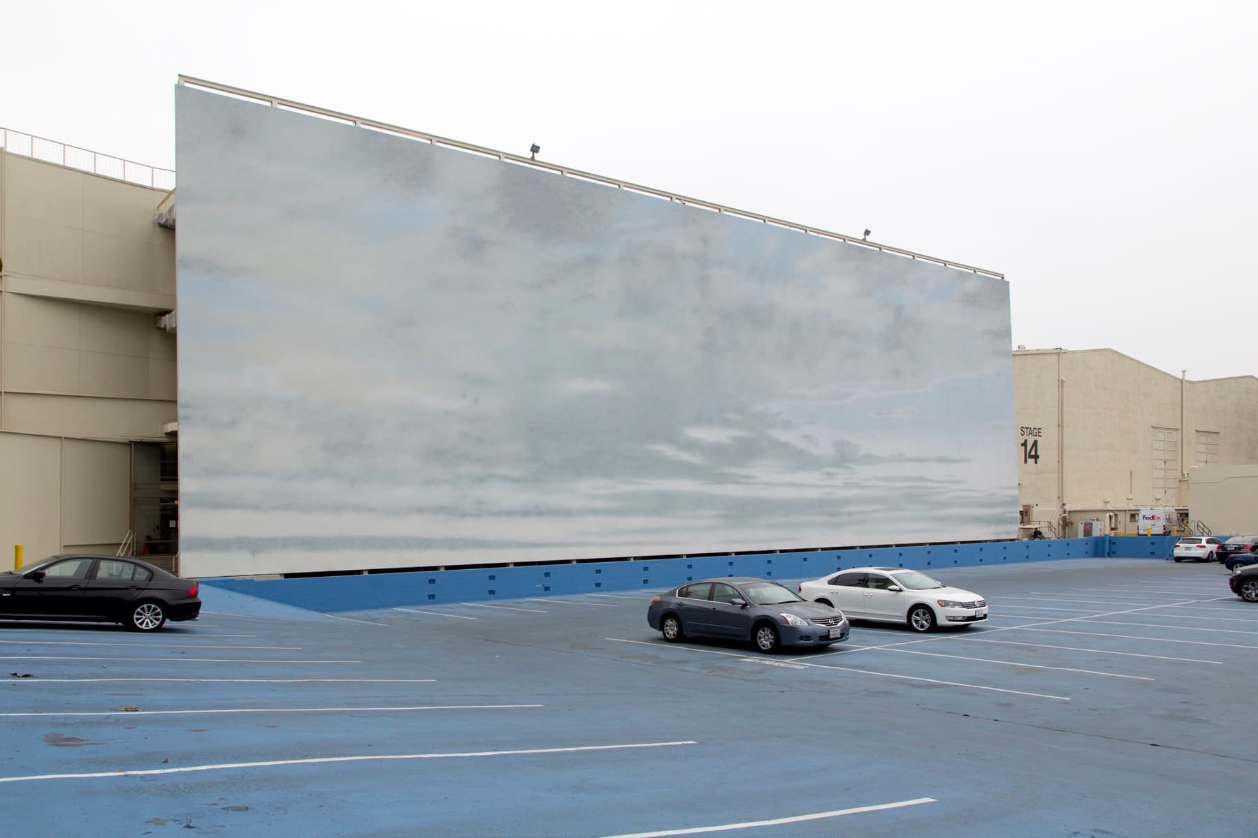 A huge flat wall painted with pale blue sky and clouds rising behind a car park with a few parked cars