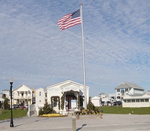 A wide lawn and paved square with a tall flagpole, the white post office and pastel houses behind