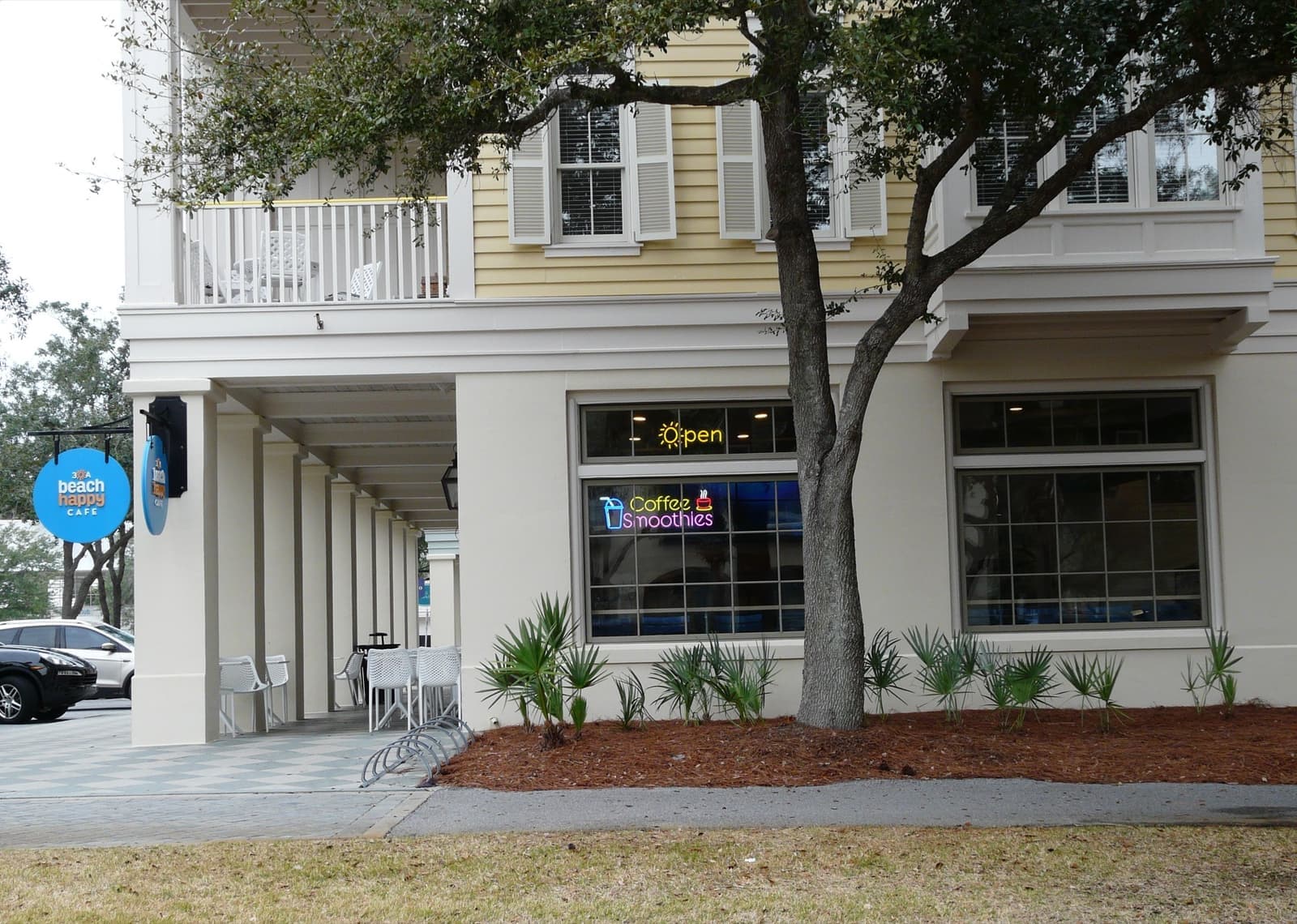 A pale yellow and white building with a colonnade and a cafe sign under an oak tree