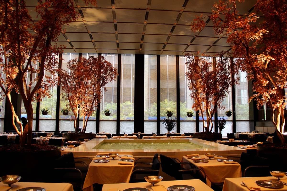 A high dining room with a square white marble pool, trees at the corners and rippling curtains