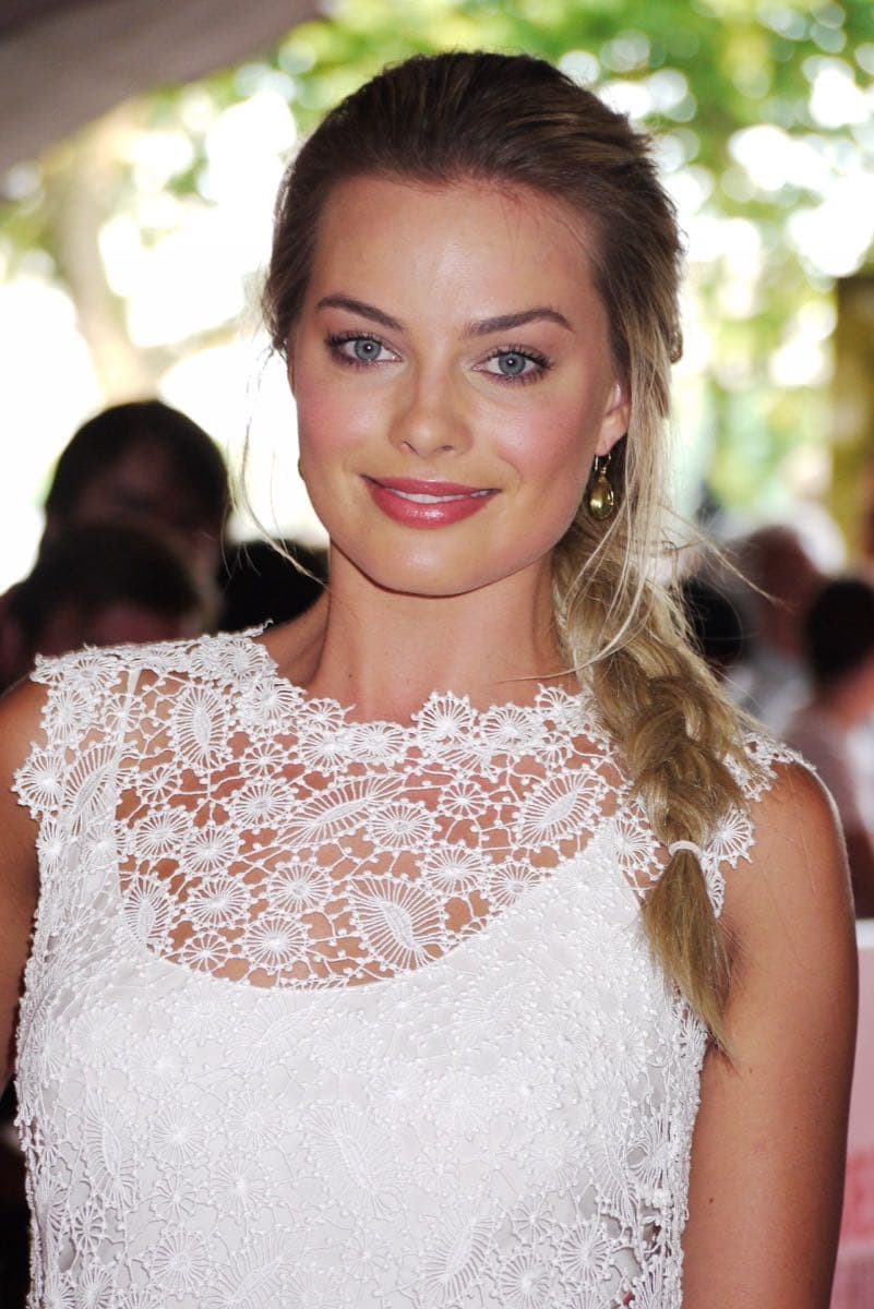 Margot Robbie in a white lace dress, her hair in a side plait, smiling