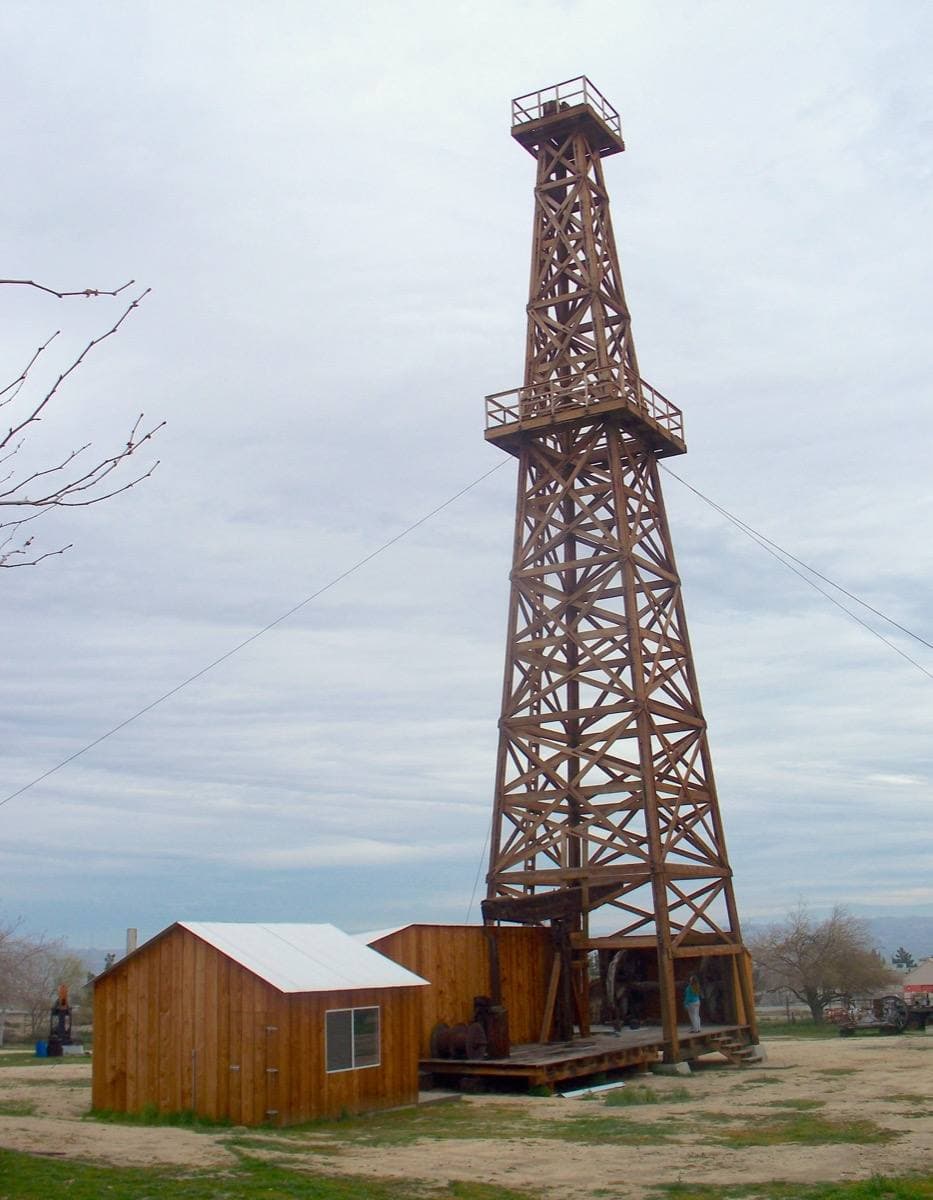 A tall timber oil derrick against a pale sky, a small shed at its foot