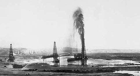 A black-and-white photograph of a column of oil shooting above a derrick into the sky