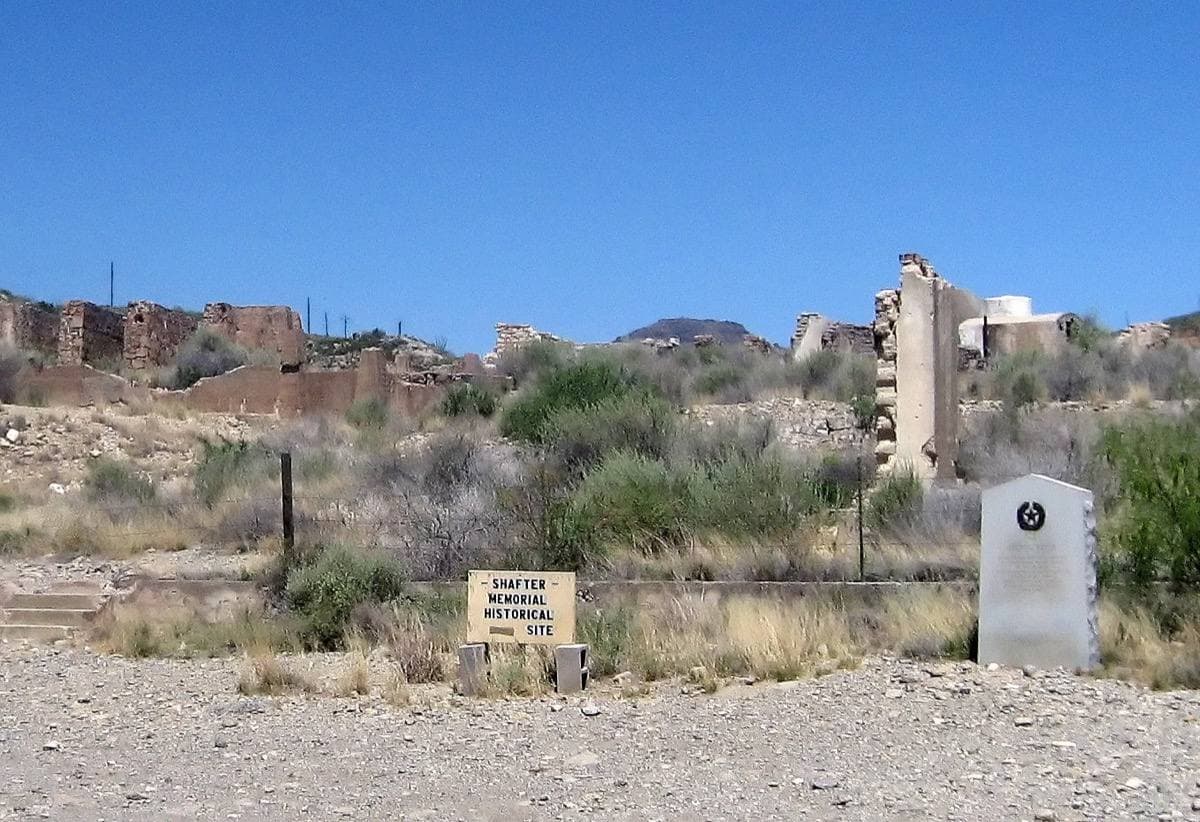 Ruined stone walls among desert scrub under a blue sky, a historical marker in front
