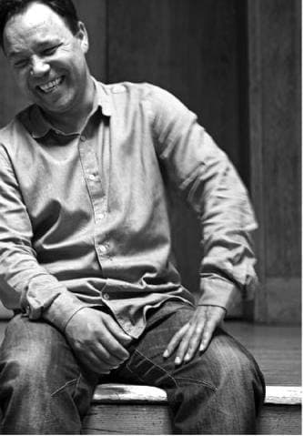Stephen Graham in a pale shirt and jeans, laughing, in black and white