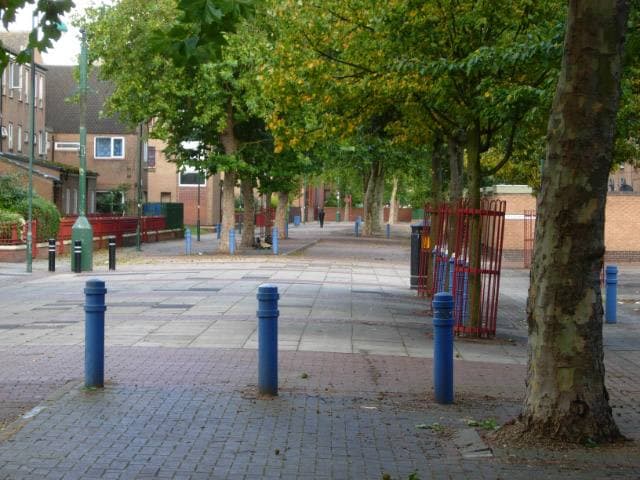 A paved walk with blue bollards and plane trees between brick houses