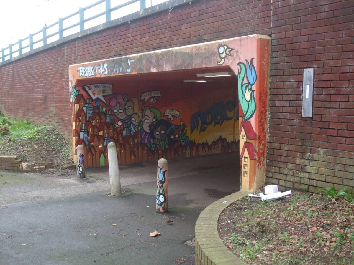 A painted pedestrian underpass under a brick embankment, with bollards in front