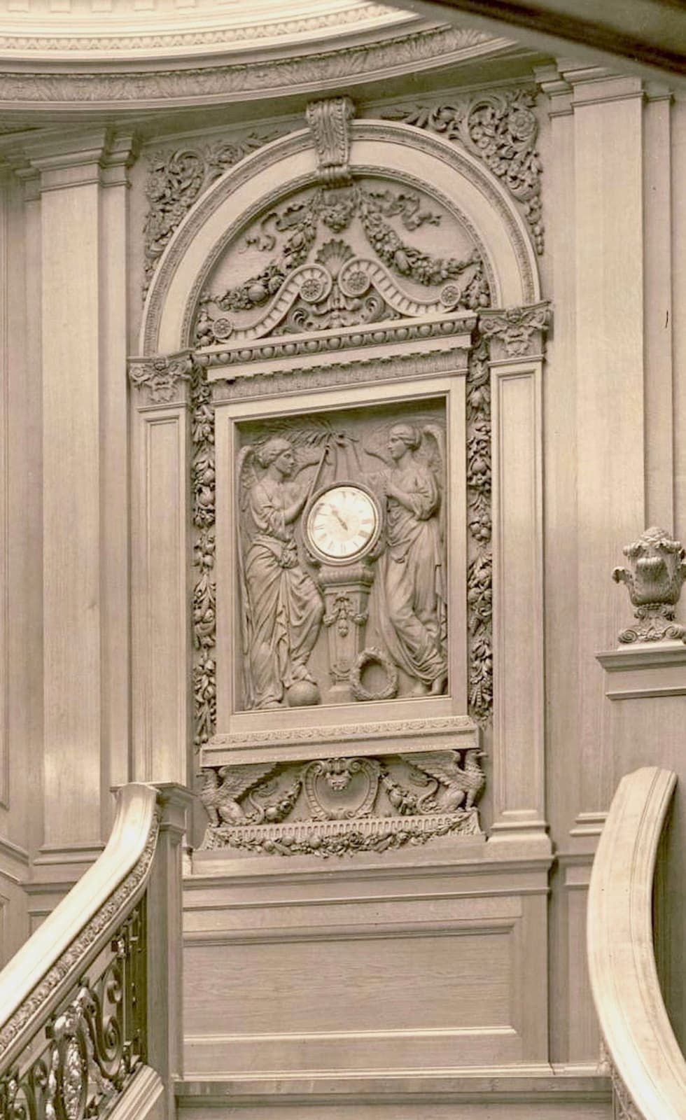 A carved panel with a clock face flanked by two allegorical figures