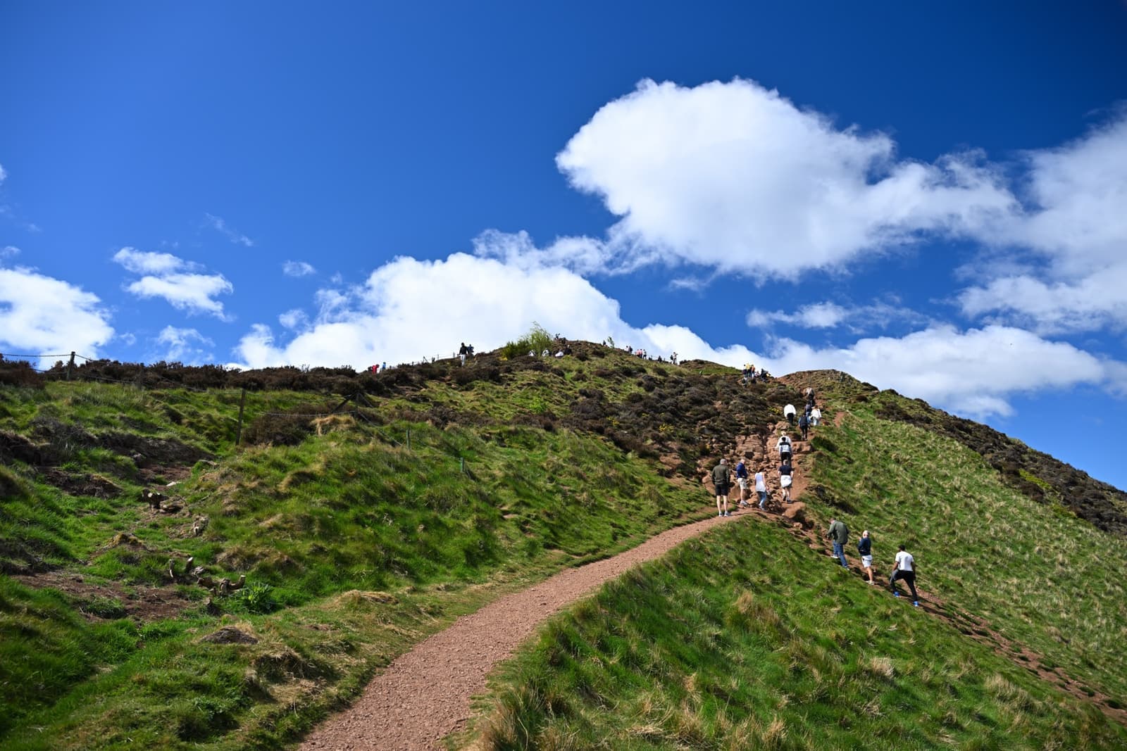 People climb a steep grassy path towards a summit under a blue sky