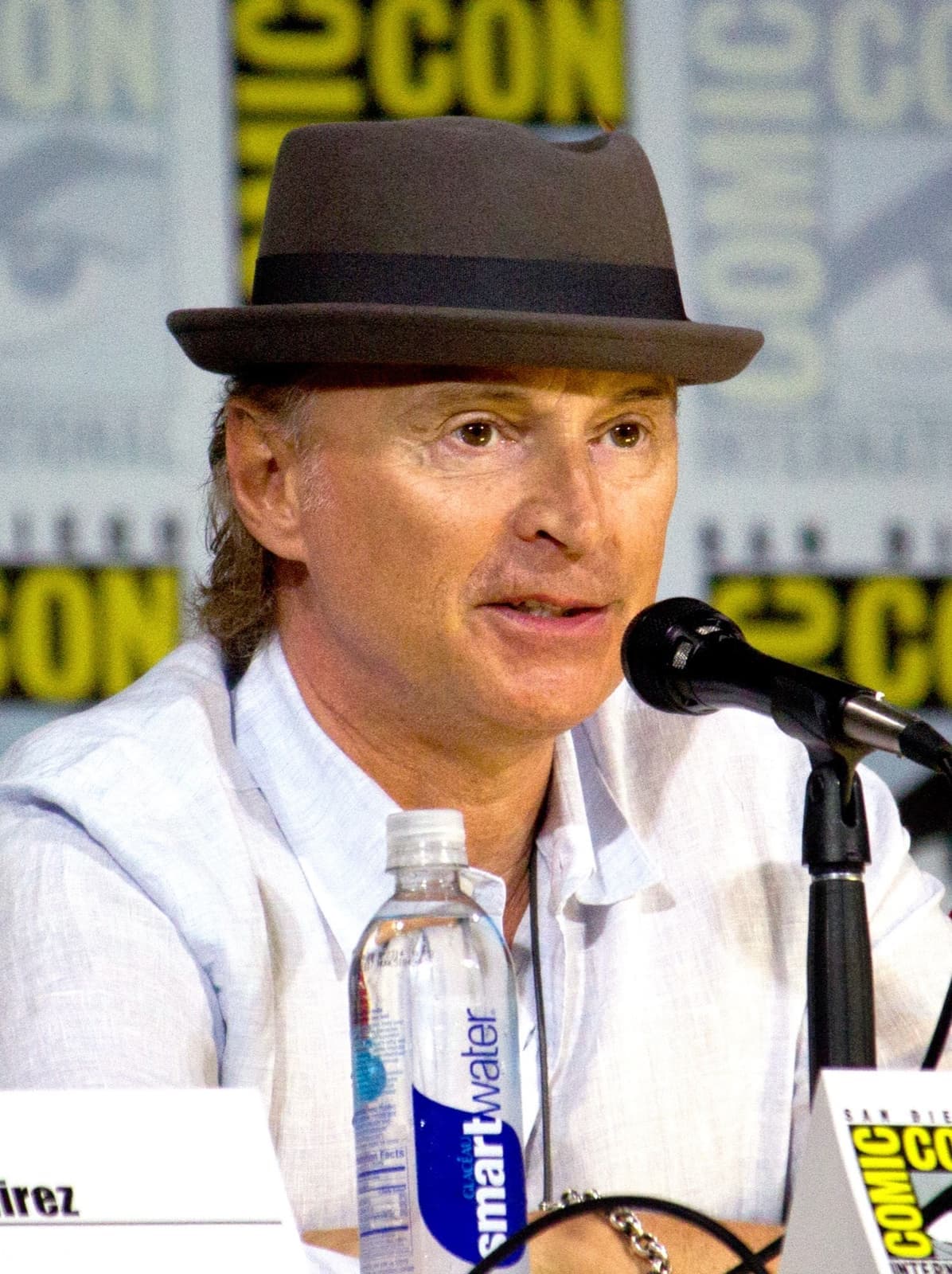 Robert Carlyle in a grey trilby and white shirt, speaking into a microphone on a panel