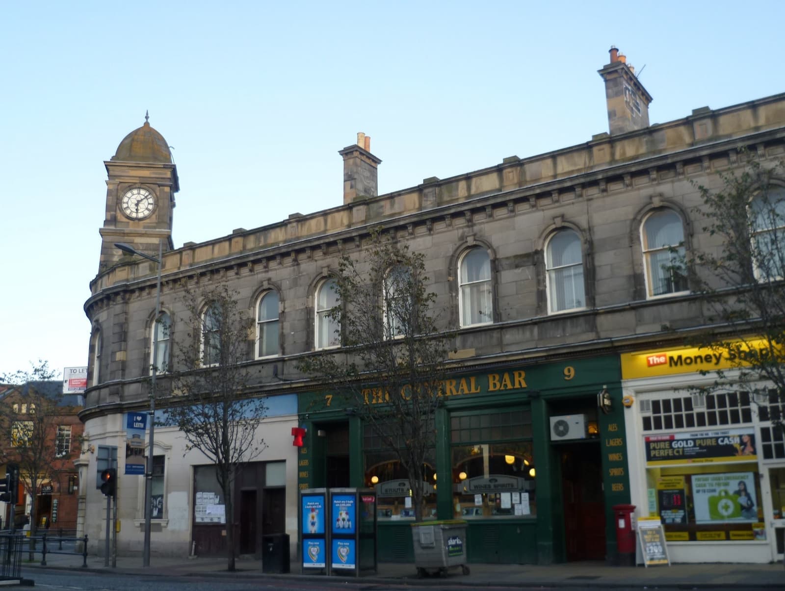 A long sandstone building with a clock tower at the corner and a green pub front