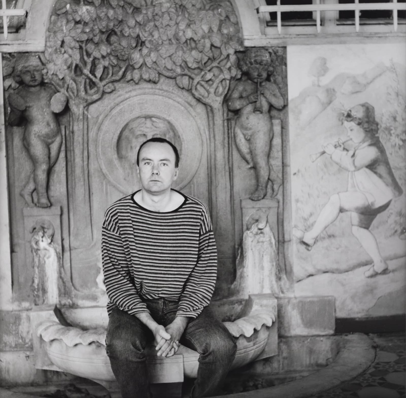 Black and white portrait of Irvine Welsh in a striped jumper, seated in front of a carved stone fountain