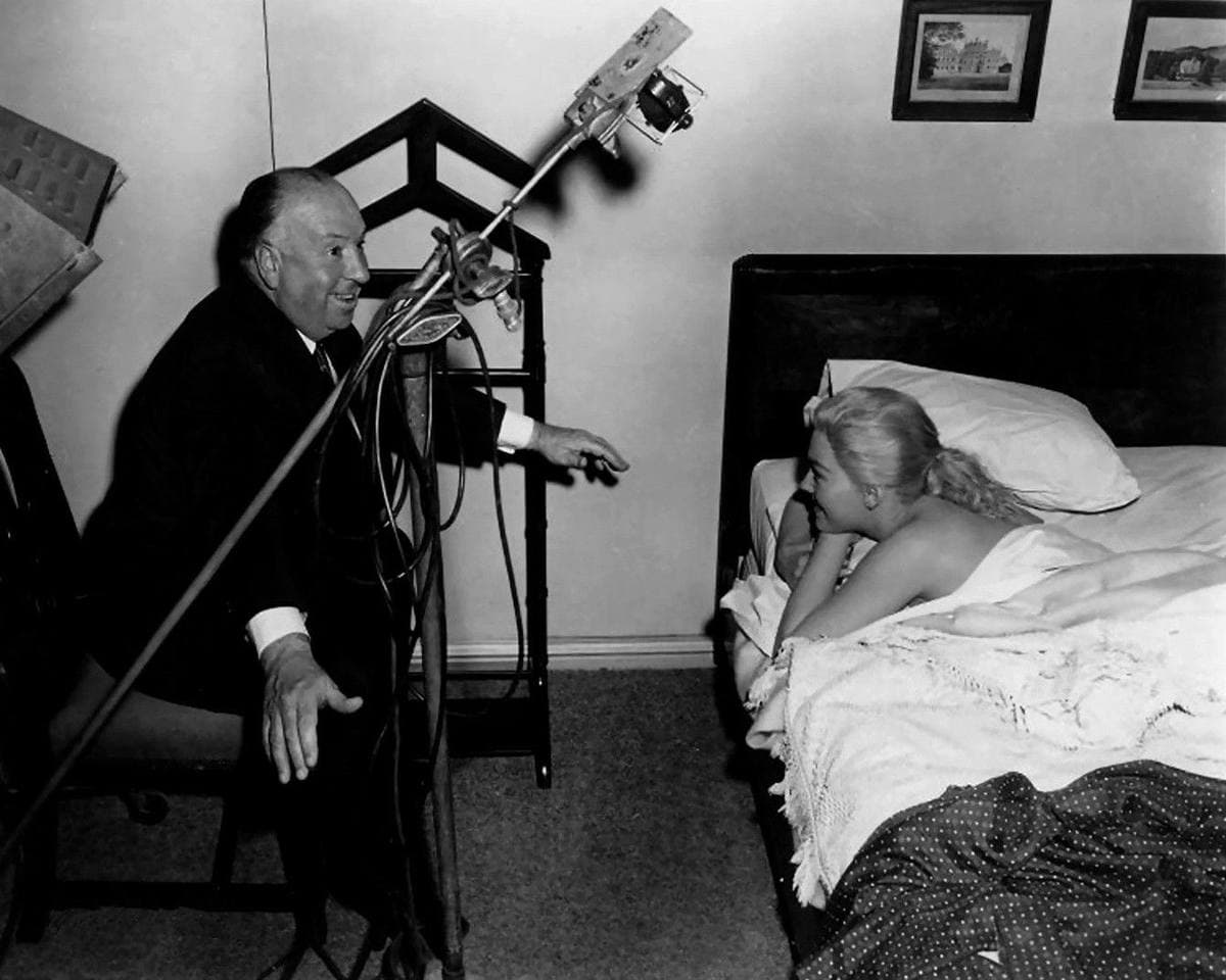 Alfred Hitchcock, seated, gesturing to Kim Novak lying on a bed on set