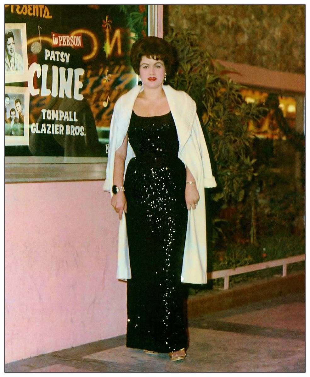 A woman in a long black sequinned dress and white stole stands by a poster bearing her name