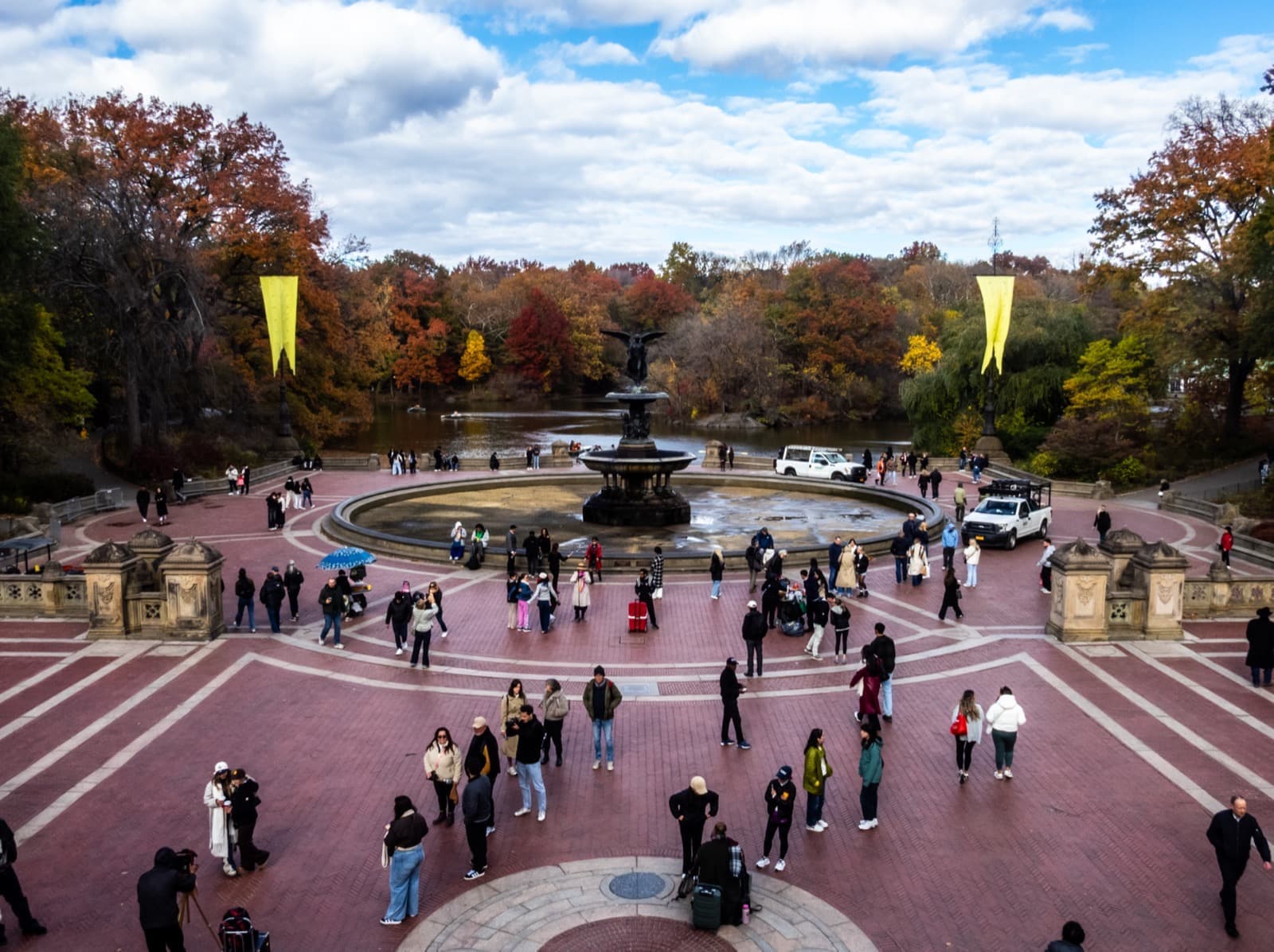 A brick-paved terrace round a fountain, ringed by autumn trees and people walking