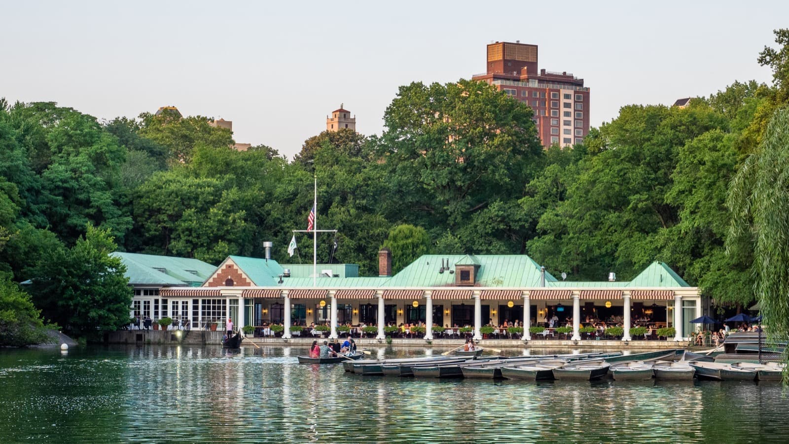 A long low boathouse with a green roof at the water’s edge, trees behind