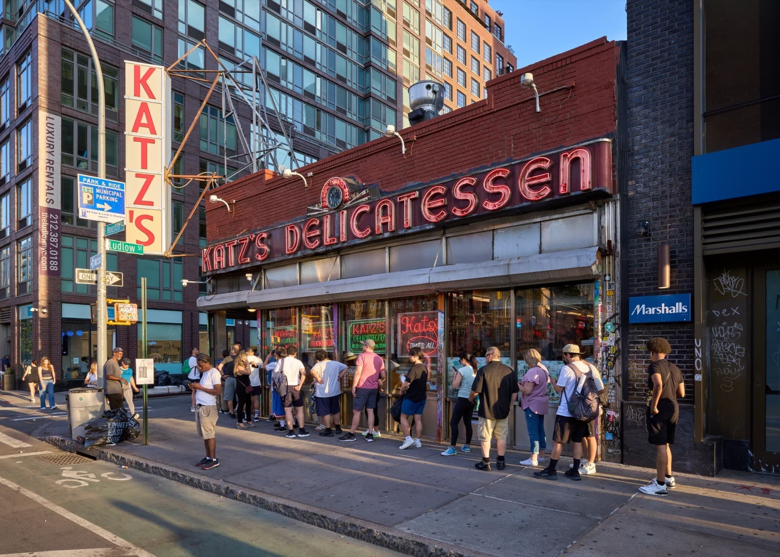 A corner deli with a long red sign and a queue along the pavement